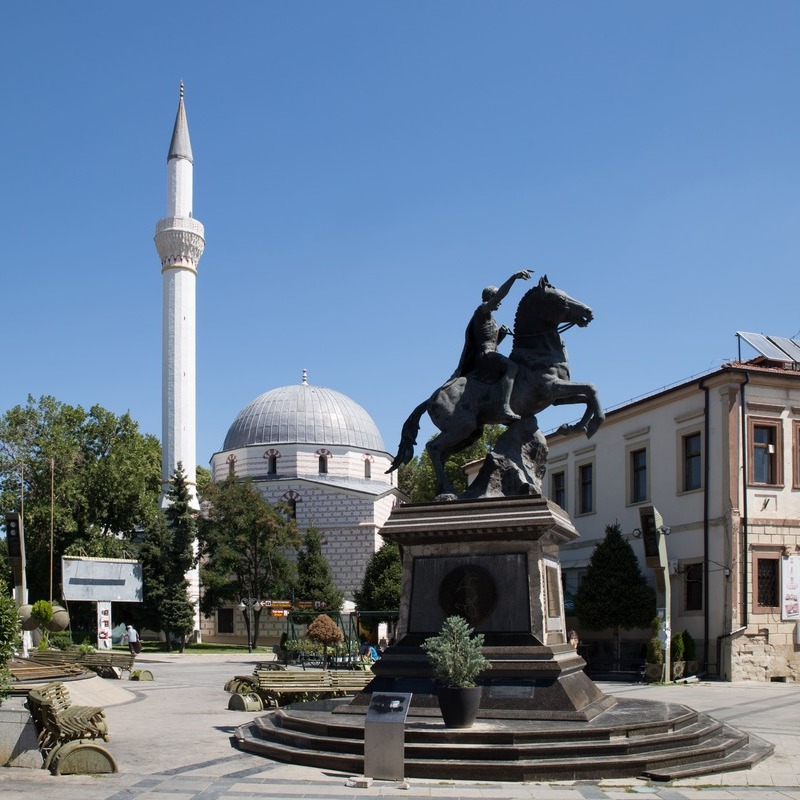 Statue Of A Warrior Riding A Horse In Central Bitola, Depicted Against The Backdrop Of A White Mosque, North Macedonia, South Eastern Europe