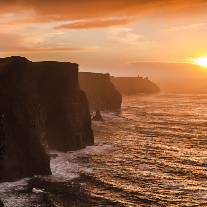 The sky above the Cliffs of Moher is illuminated orange as the sun rests over the water