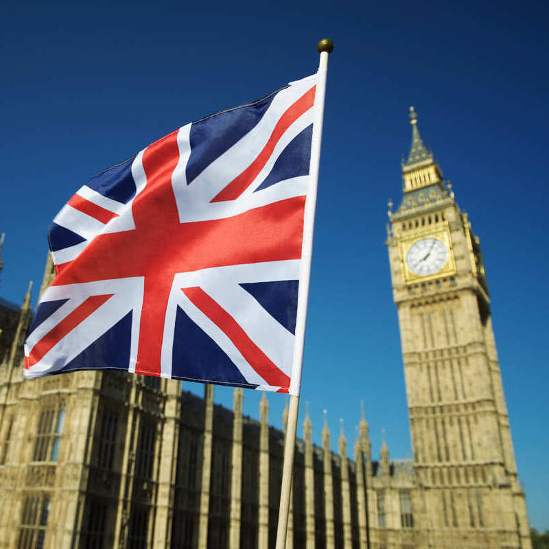 Union Jack Being Waved In Front Of Westminster Palace, Elizabeth Tower With Big Ben Clock Appearing Blurred In The Background, London, England, United Kingdom