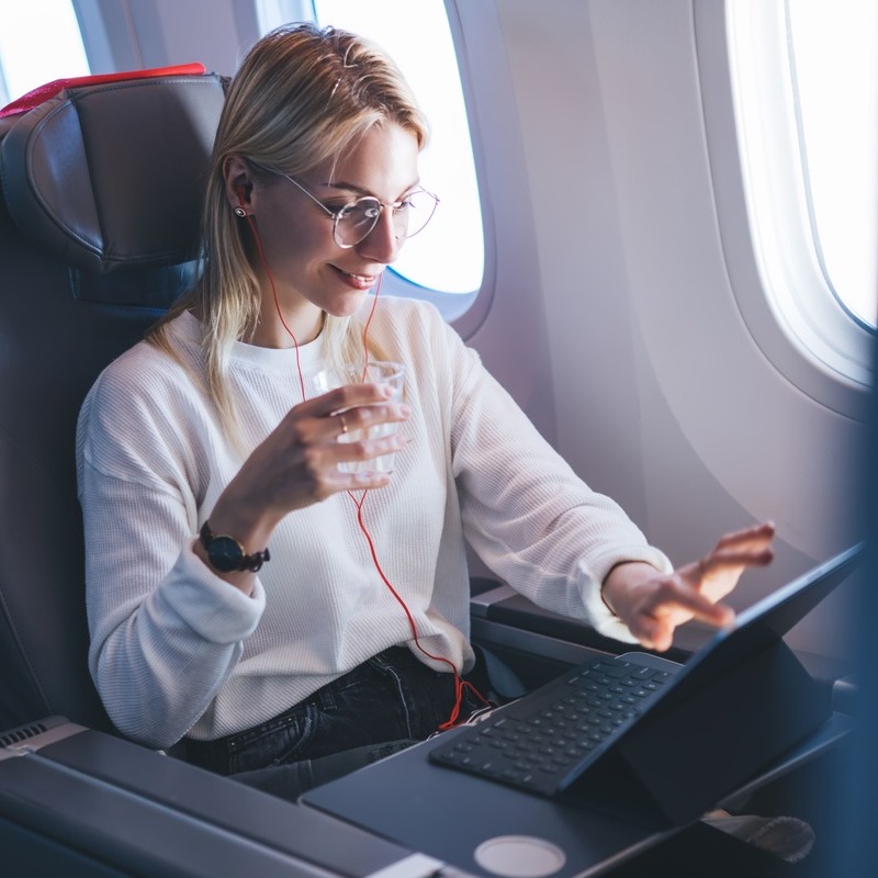 Young Female Traveler Smiling As She Works From Her Computer While Listening To Music Inside An Aircraft, International Travel