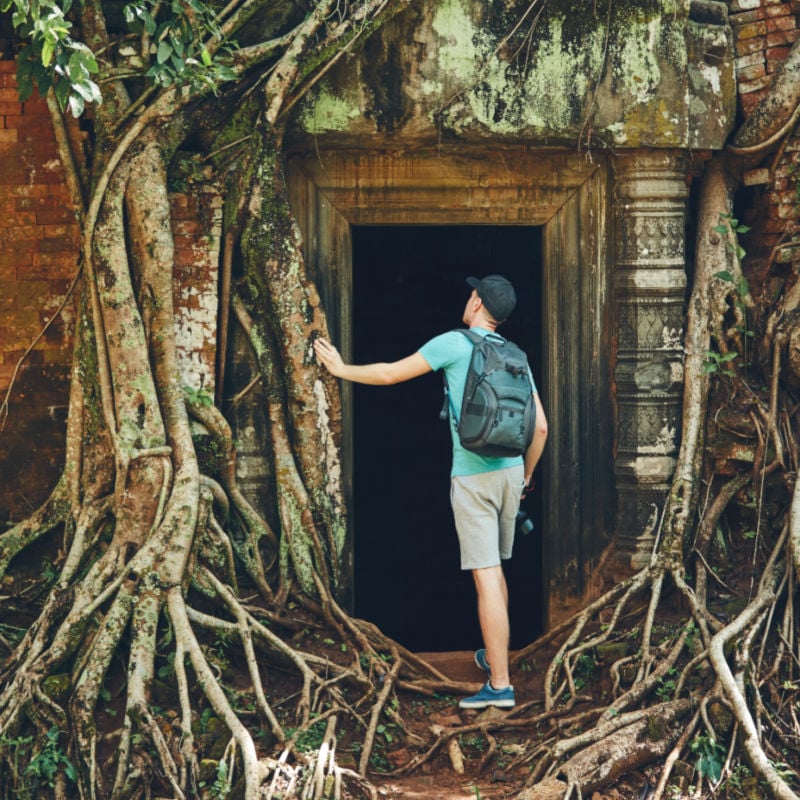 Young man with backpack coming to ancient monuments under the giant roots of the tree near Siem Reap (Angkor Wat) in Cambodia