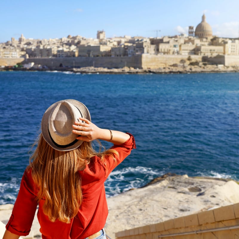 Beautiful young woman with hat descends stairs in Malta towards waterfront looking at panoramic view of Valletta, Malta copy