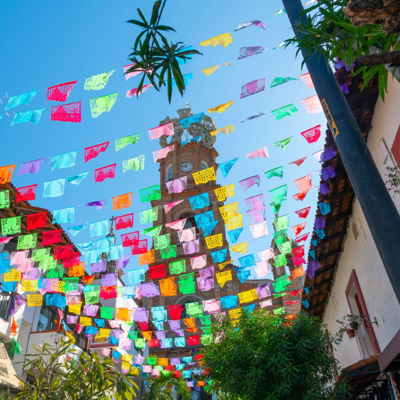 View from the street below of the The Church of Our Lady of Guadalupe with colorful flags strung across the street on a sunny morning in Puerto Vallarta