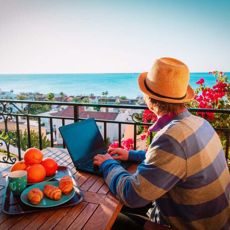 Male Remote Worker Or Digital Nomad Wearing Summery Clothes As He Works From A Balcony In A Coastal Location With Some Fruit And Croissant On The Table, Spain